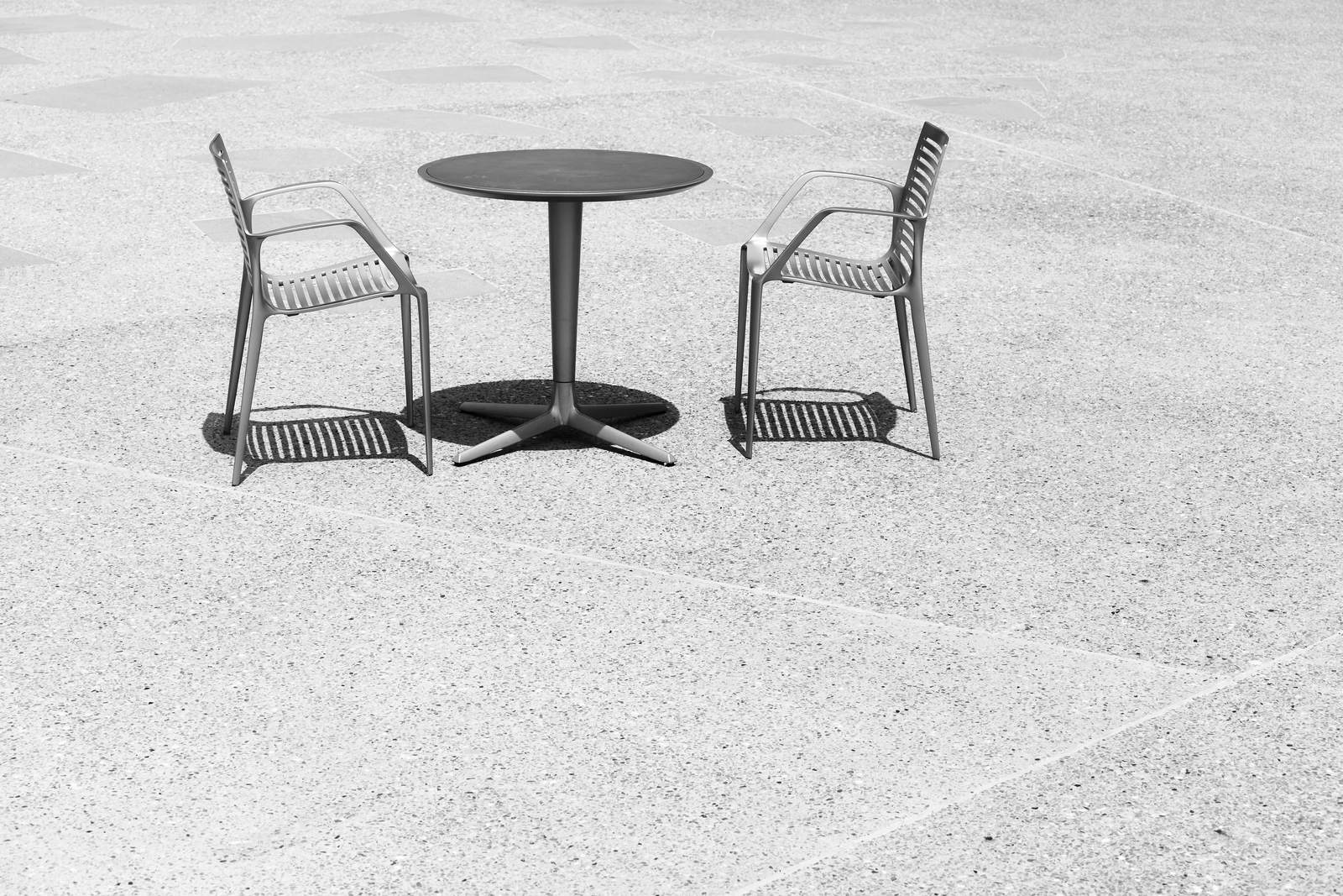 Round outdoor table and two chairs casting shadows on a gray concrete patio